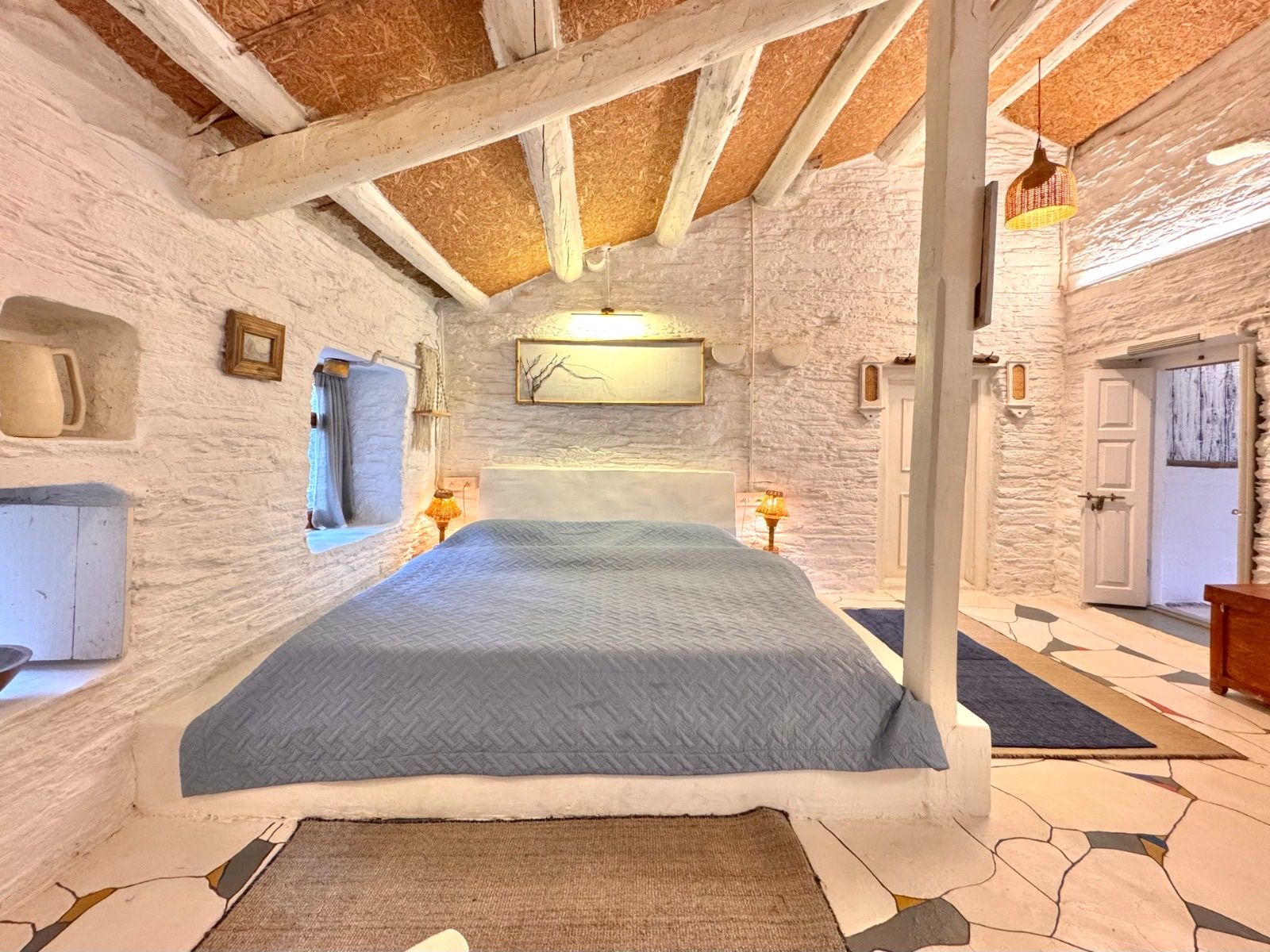 A bedroom in Wabi and Sabi, with whitewashed stone walls, white beams and a blue bedcover